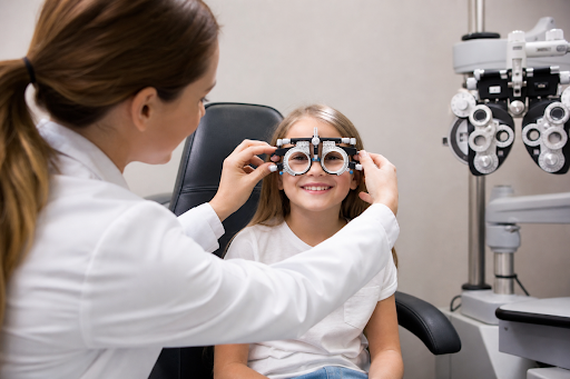 Optometrist fits trial frame glasses on a smiling child during an eye exam, phoropter beside them.